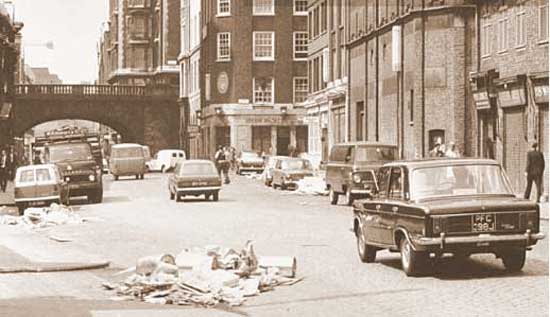 Lower Thames Street 1970, looking W towards London Bridge, before  demolition of buildings on N side of the street.  Joe’s Café  is at the corner of Fish Street Hill, with a Mini parked outside. The rubbish in the street from the Billingsgate early morning fish market was collected at lunch time.